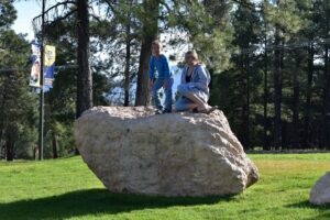 Two people sitting on a large rock in the grass.