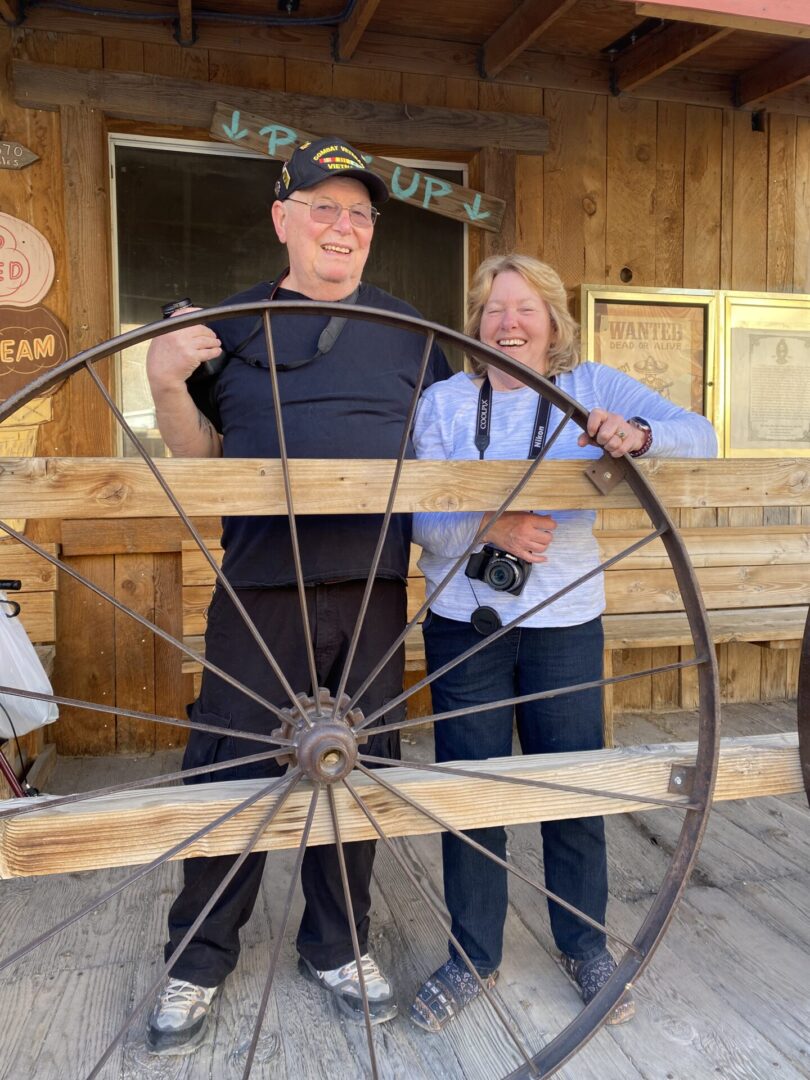 A man and woman standing next to an old wagon wheel.