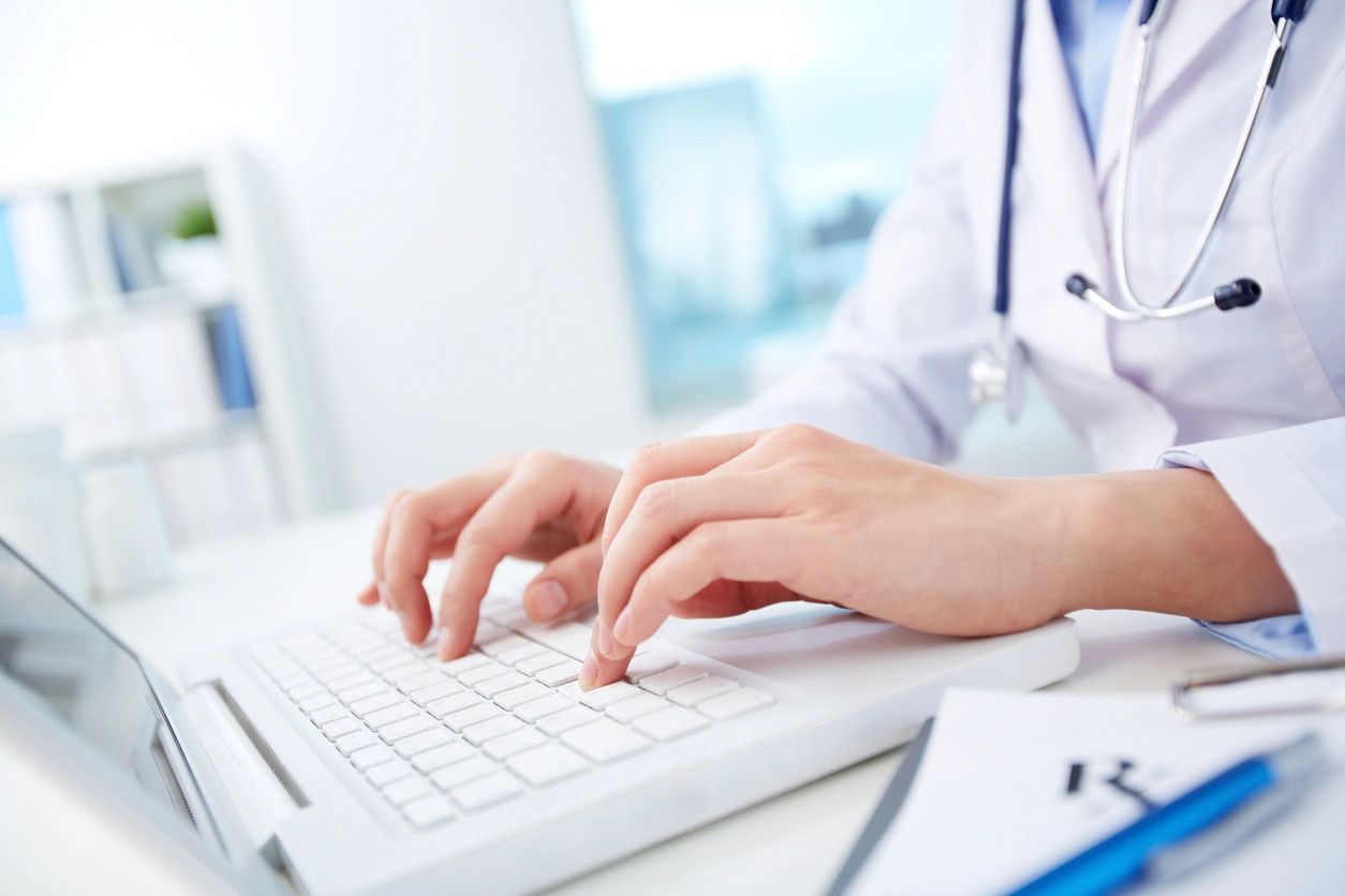 A person typing on a laptop with a stethoscope around their neck.